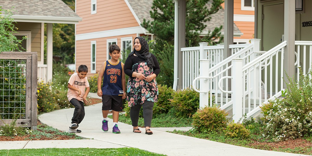 A woman and two boys walk along a pathway in a residential neighborhood, surrounded by greenery and colorful homes.