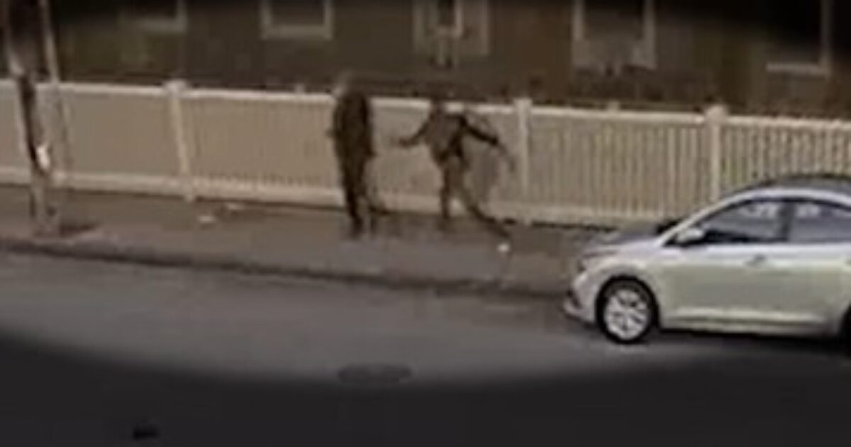 Two individuals walking on a sidewalk near a white car, with a fence in the background.