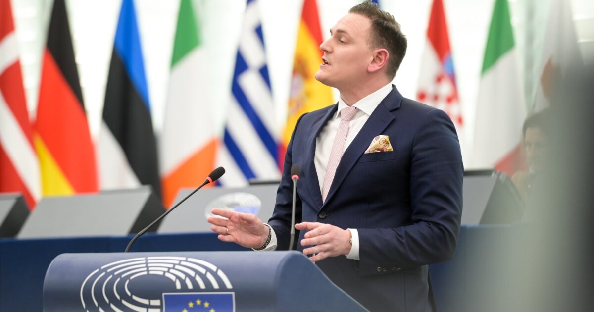 Man in a suit speaks at the European Parliament, with various national flags in the background and a podium displaying the European Union emblem.