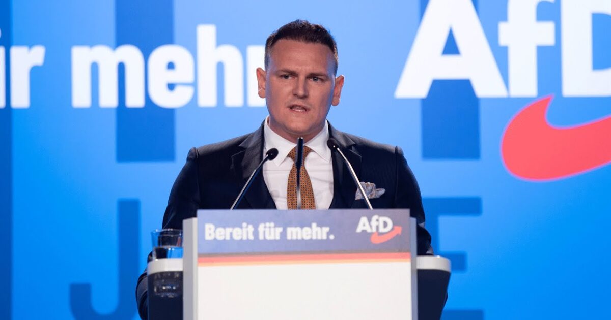 A speaker addresses an audience at an AfD event, emphasizing key political messages with a backdrop featuring the party's branding.