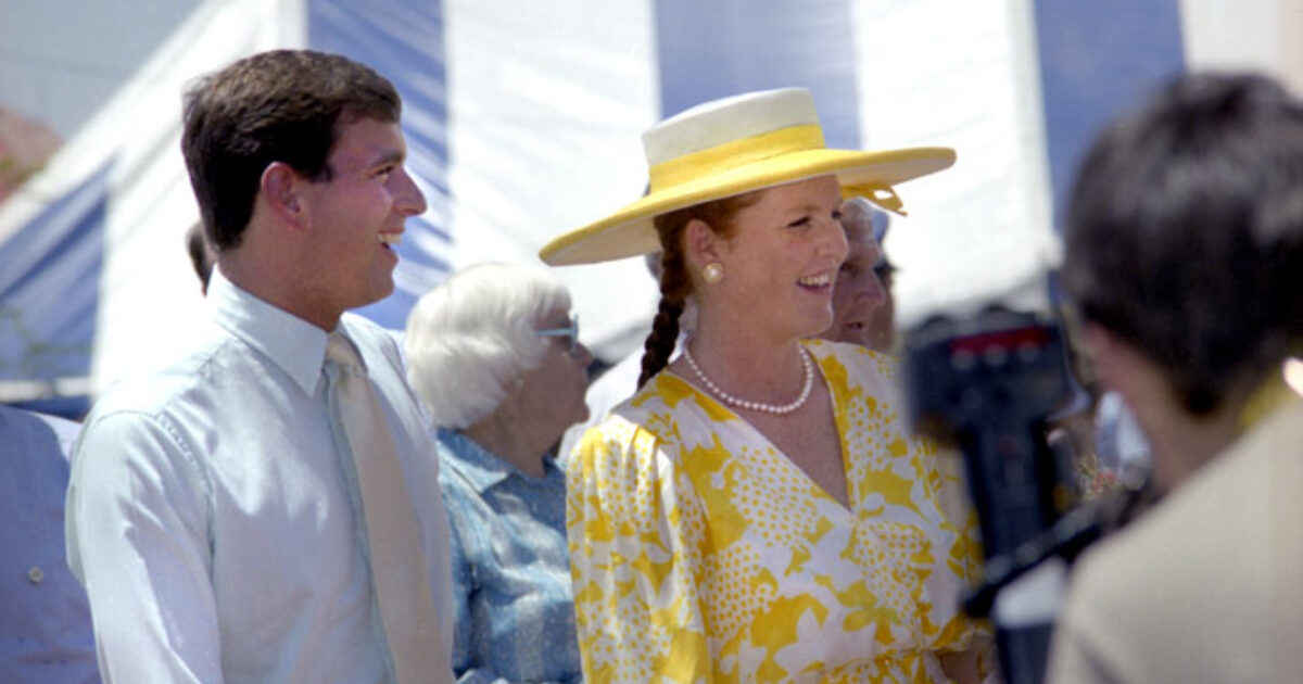 A smiling woman in a yellow floral dress and wide-brimmed hat stands beside a man in a light blue shirt, both attending a festive outdoor event.