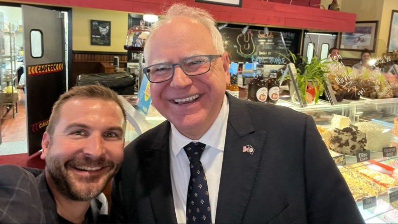 Selfie of two men smiling in a café with desserts in the background, showcasing a friendly atmosphere and community engagement.