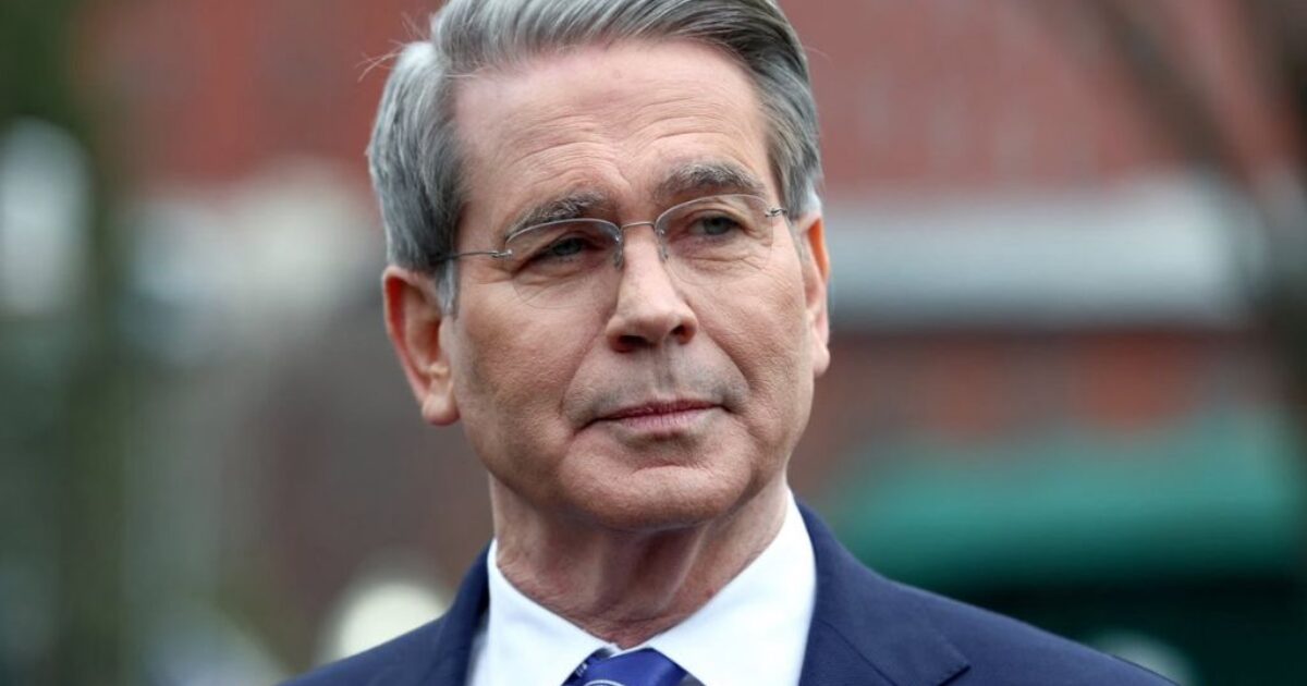 Man in a suit with gray hair and glasses, looking thoughtfully while standing outdoors against a blurred background.