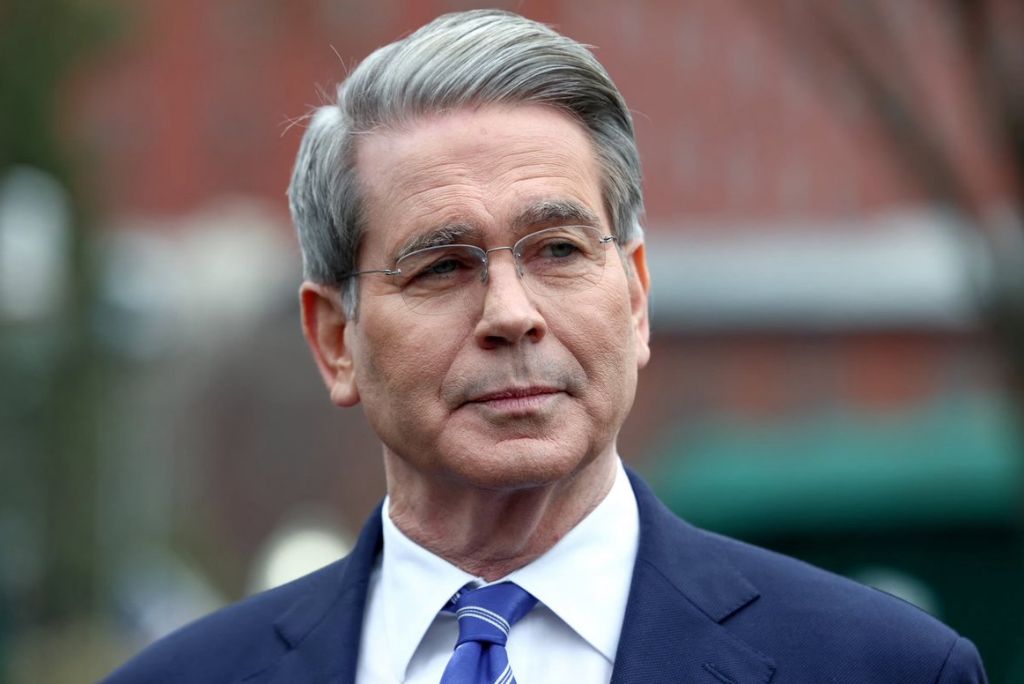 Man in a suit with gray hair and glasses, looking thoughtfully while standing outdoors against a blurred background.