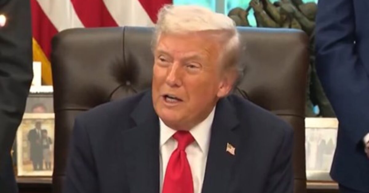 Donald Trump seated in the Oval Office, wearing a suit and red tie, with American flags and historical artifacts in the background.