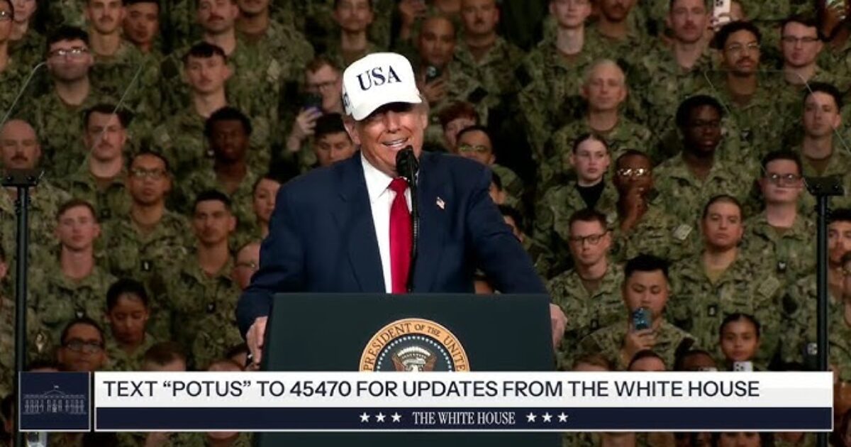 Former President Donald Trump speaks at a military event wearing a "USA" hat, addressing a crowd of service members.