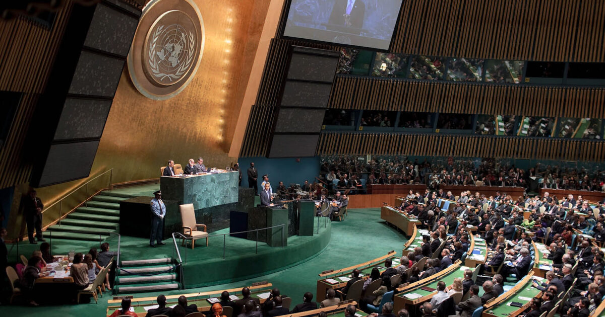 United Nations General Assembly session with delegates and officials engaged in discussion, featuring the iconic UN emblem in the background.