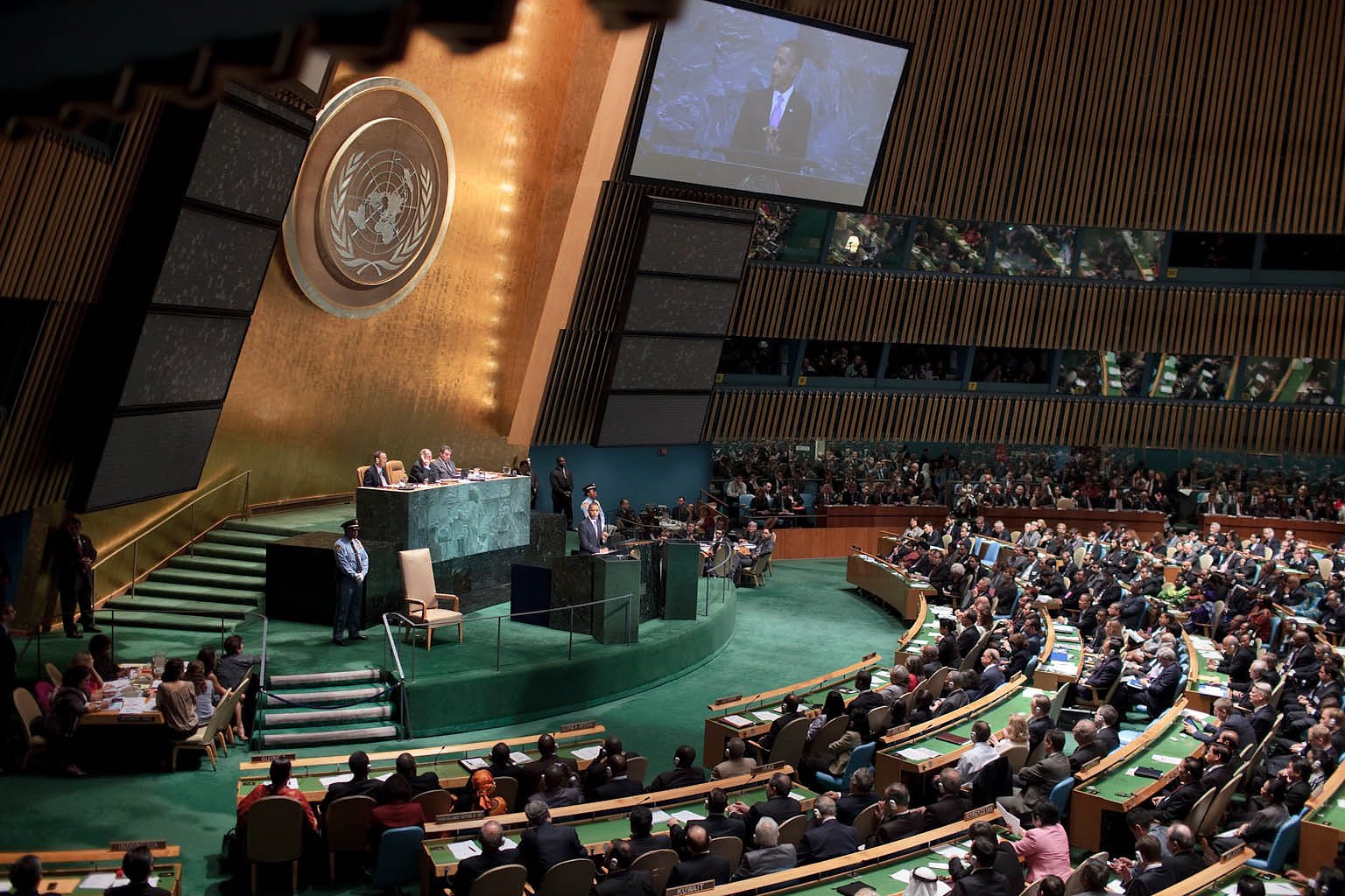 United Nations General Assembly session with delegates and officials engaged in discussion, featuring the iconic UN emblem in the background.