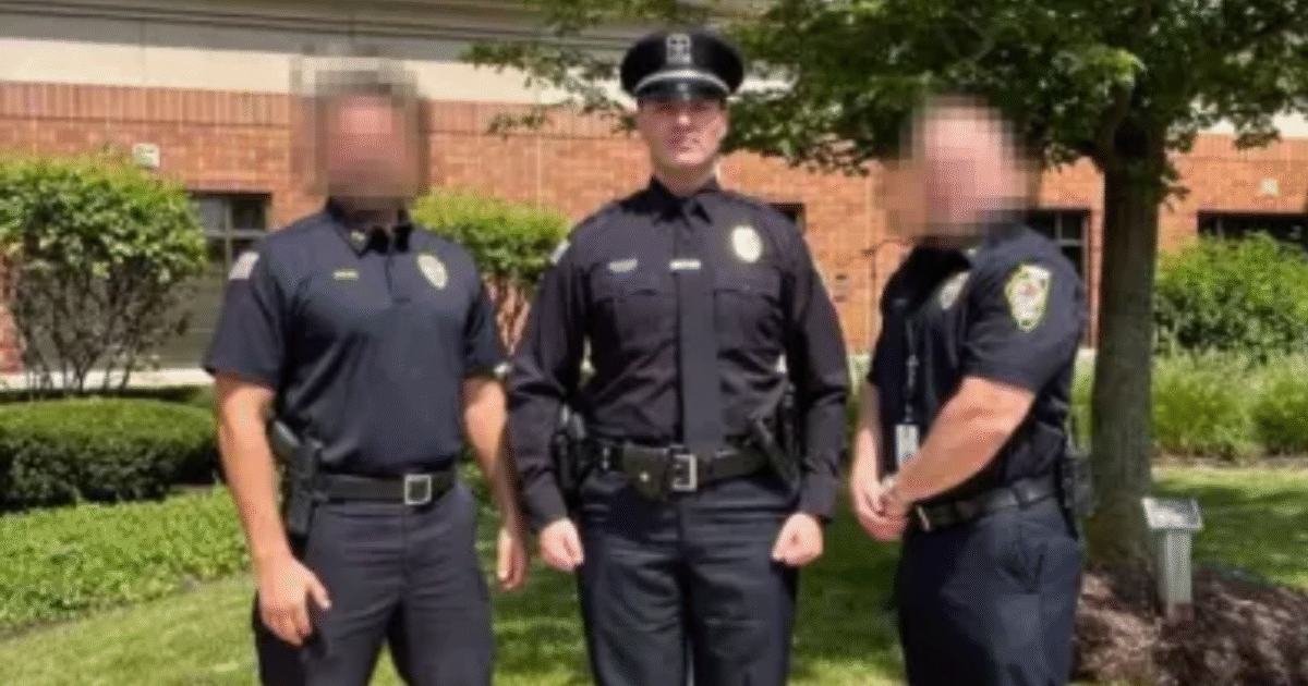 Three police officers in uniform standing together outdoors in front of a brick building surrounded by greenery.