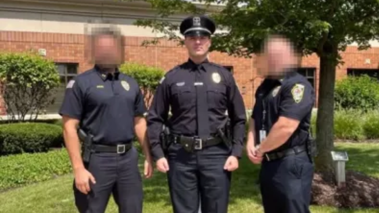 Three police officers in uniform standing together outdoors in front of a brick building surrounded by greenery.