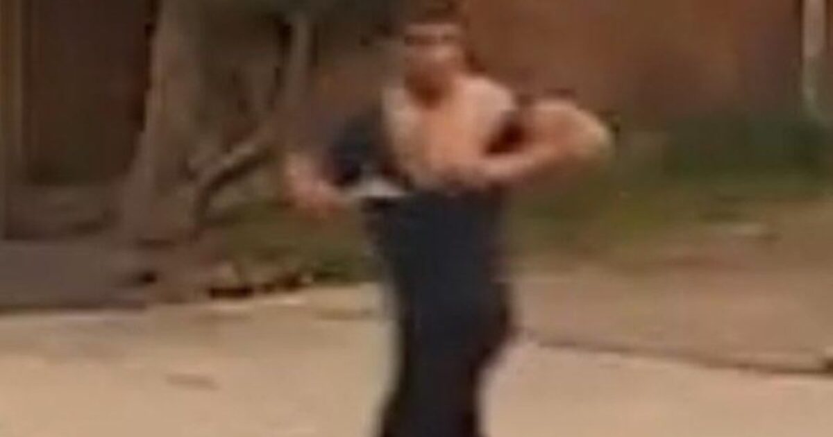 A person running on a street with a partially removed shirt, surrounded by greenery and a wooden fence in the background.