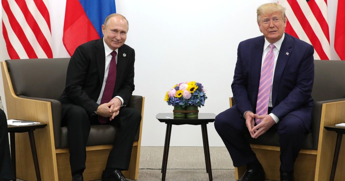 Russian President Vladimir Putin and former U.S. President Donald Trump meet for a discussion, with national flags in the background and a floral arrangement on the table.