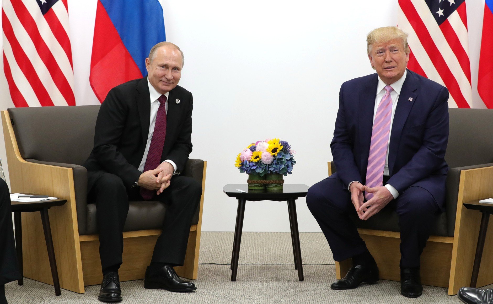 Russian President Vladimir Putin and former U.S. President Donald Trump meet for a discussion, with national flags in the background and a floral arrangement on the table.