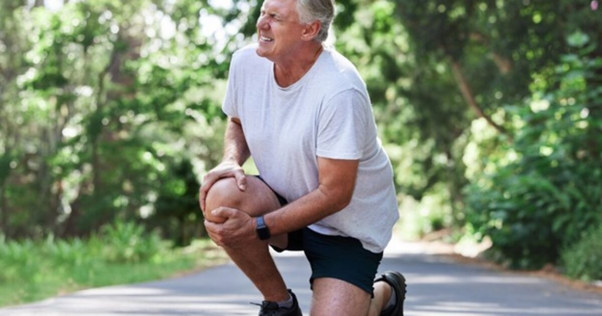 Older man experiencing knee pain while jogging on a scenic path surrounded by greenery.