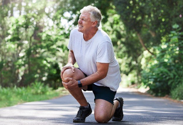 Older man experiencing knee pain while jogging on a scenic path surrounded by greenery.