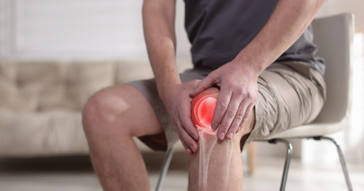 Man sitting on a chair, holding his knee in pain, indicating a potential knee injury or discomfort in a bright indoor setting.