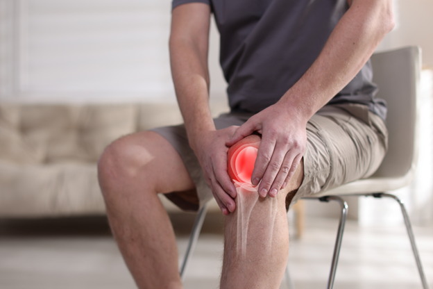 Man sitting on a chair, holding his knee in pain, indicating a potential knee injury or discomfort in a bright indoor setting.