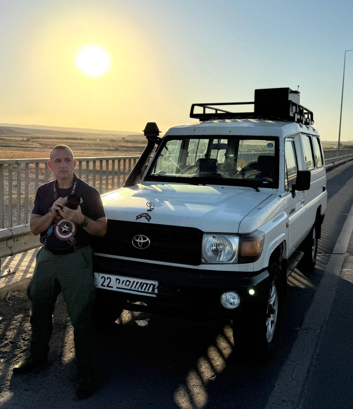 A man stands beside a white Toyota Land Cruiser at sunset, holding a camera, with a scenic landscape in the background.