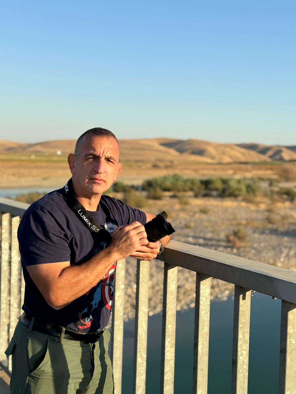 Man holding a camera while standing on a bridge with a scenic landscape of hills and a river in the background.