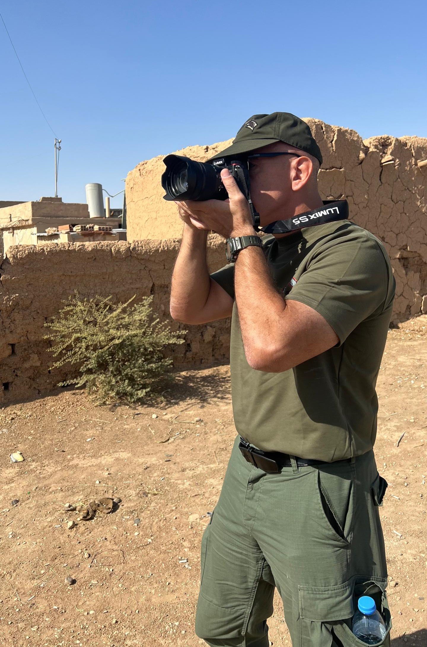 A man in a green uniform photographs a rural landscape with a DSLR camera, surrounded by mud-brick buildings under a clear blue sky.