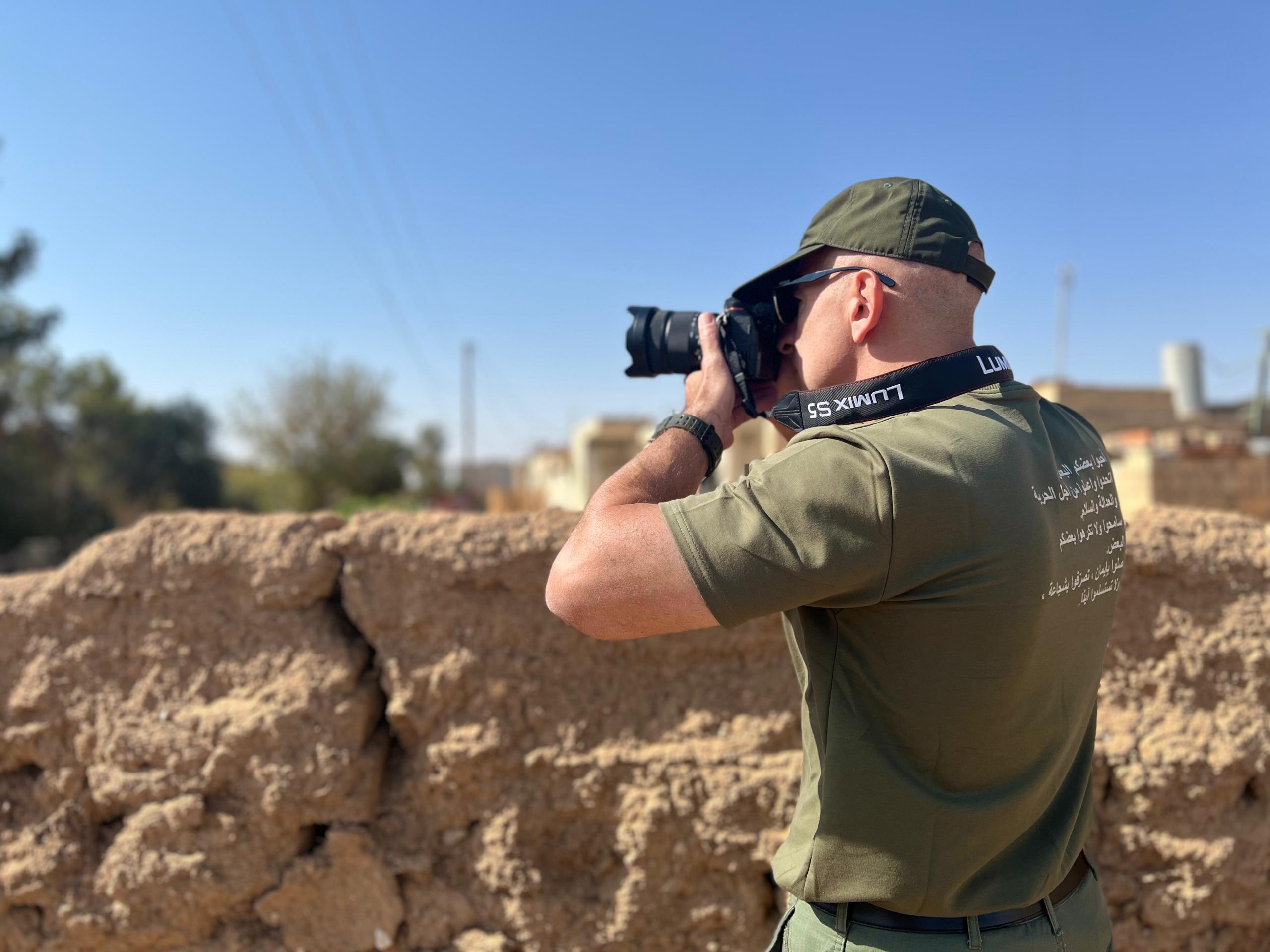 Photographer in a green shirt capturing images of a rural landscape, with a stone wall in the foreground and clear blue sky above.