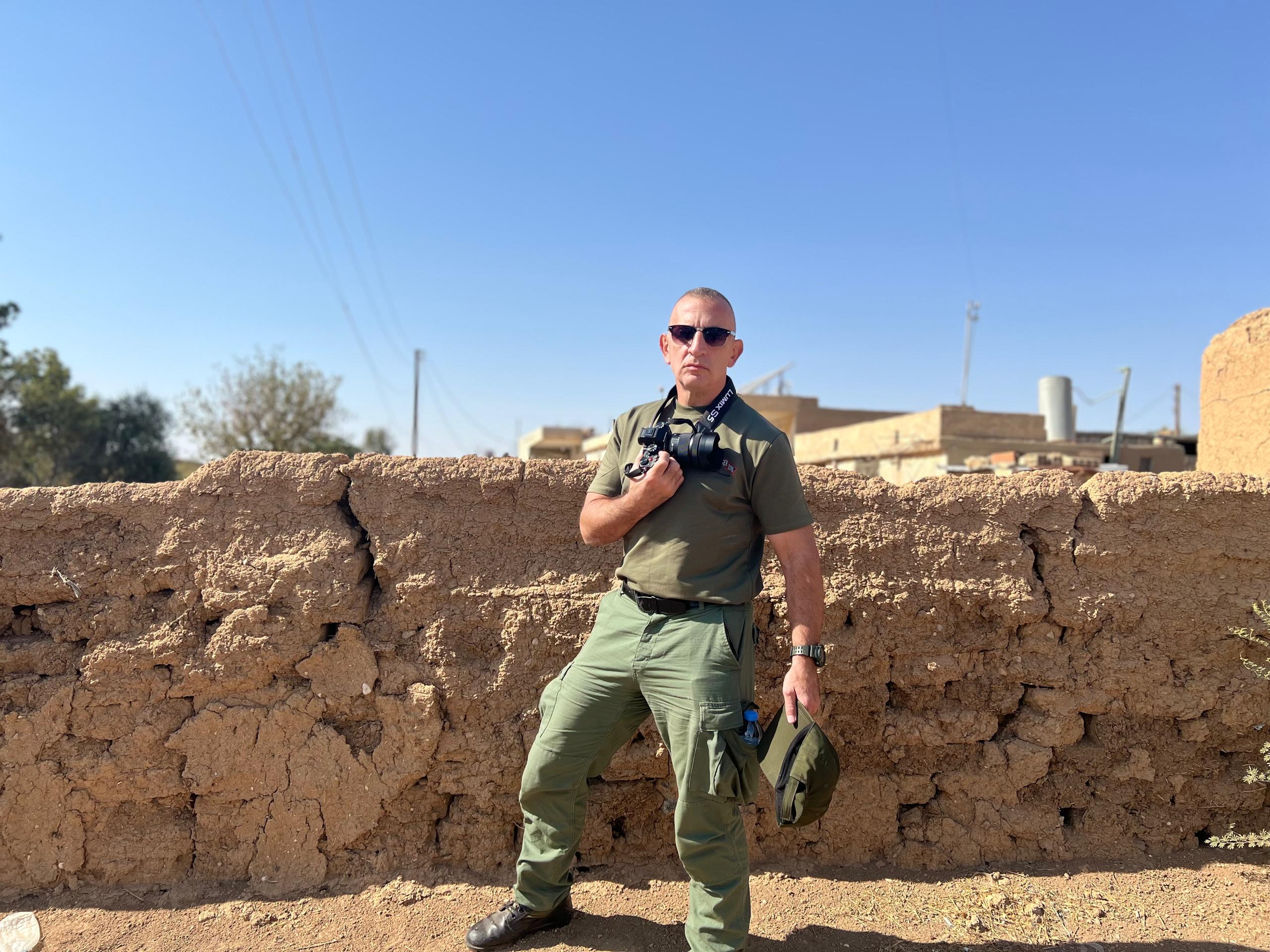 Person in military-style clothing stands against a textured earthen wall, holding a camera, with a clear blue sky in the background.