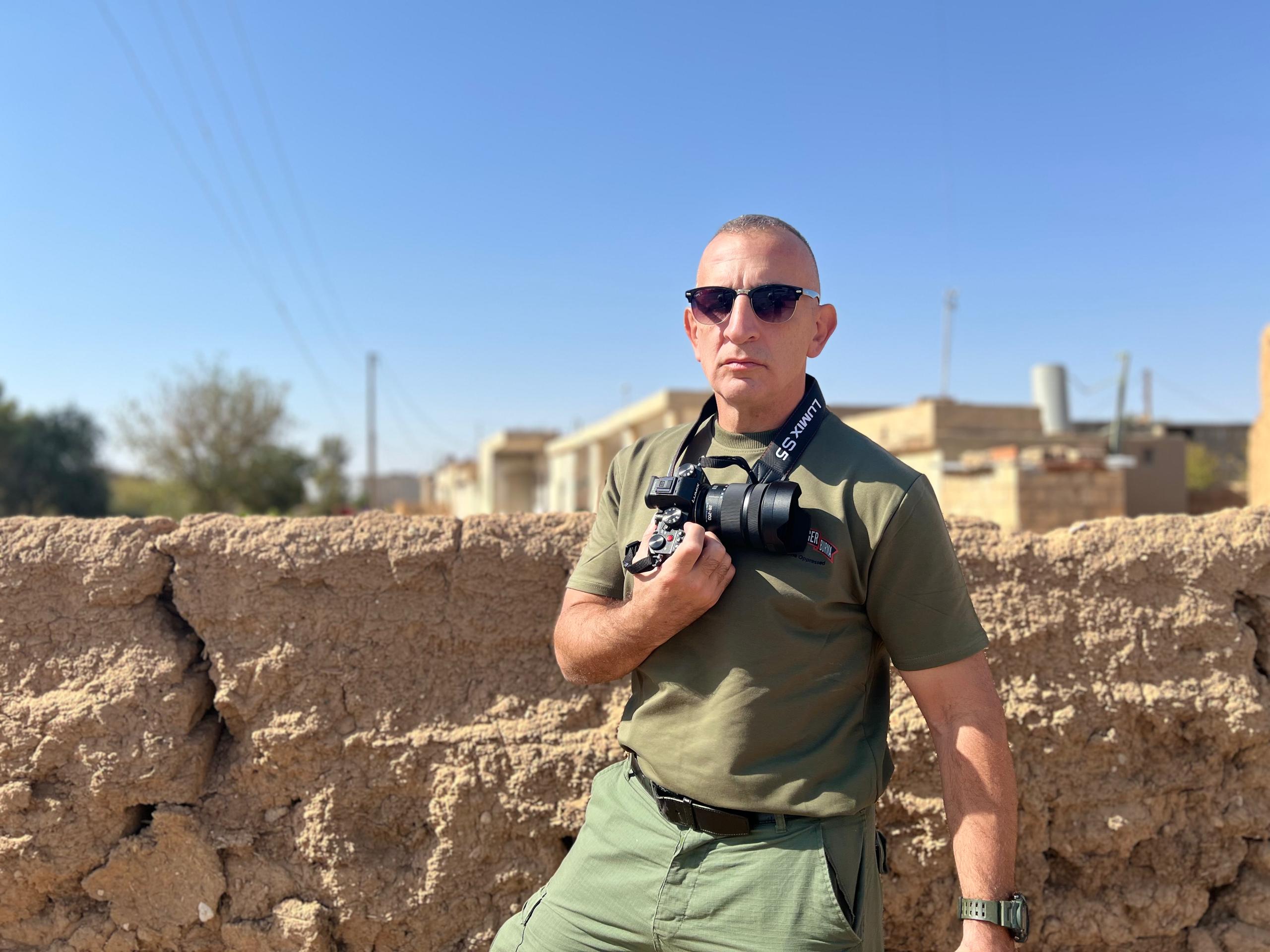 A photographer in sunglasses holds a camera while posing against a rustic wall in a sunny outdoor setting.