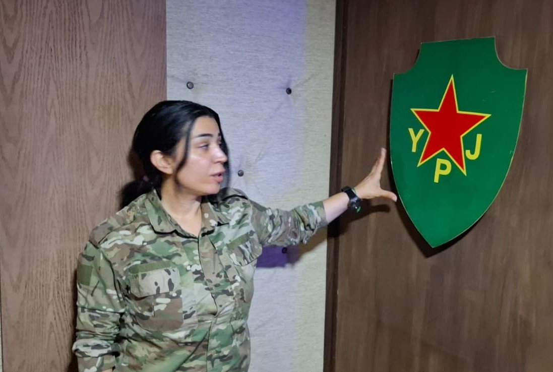 Woman in military camouflage uniform points to the YPJ emblem on a door, symbolizing the Women's Protection Units in a military setting.