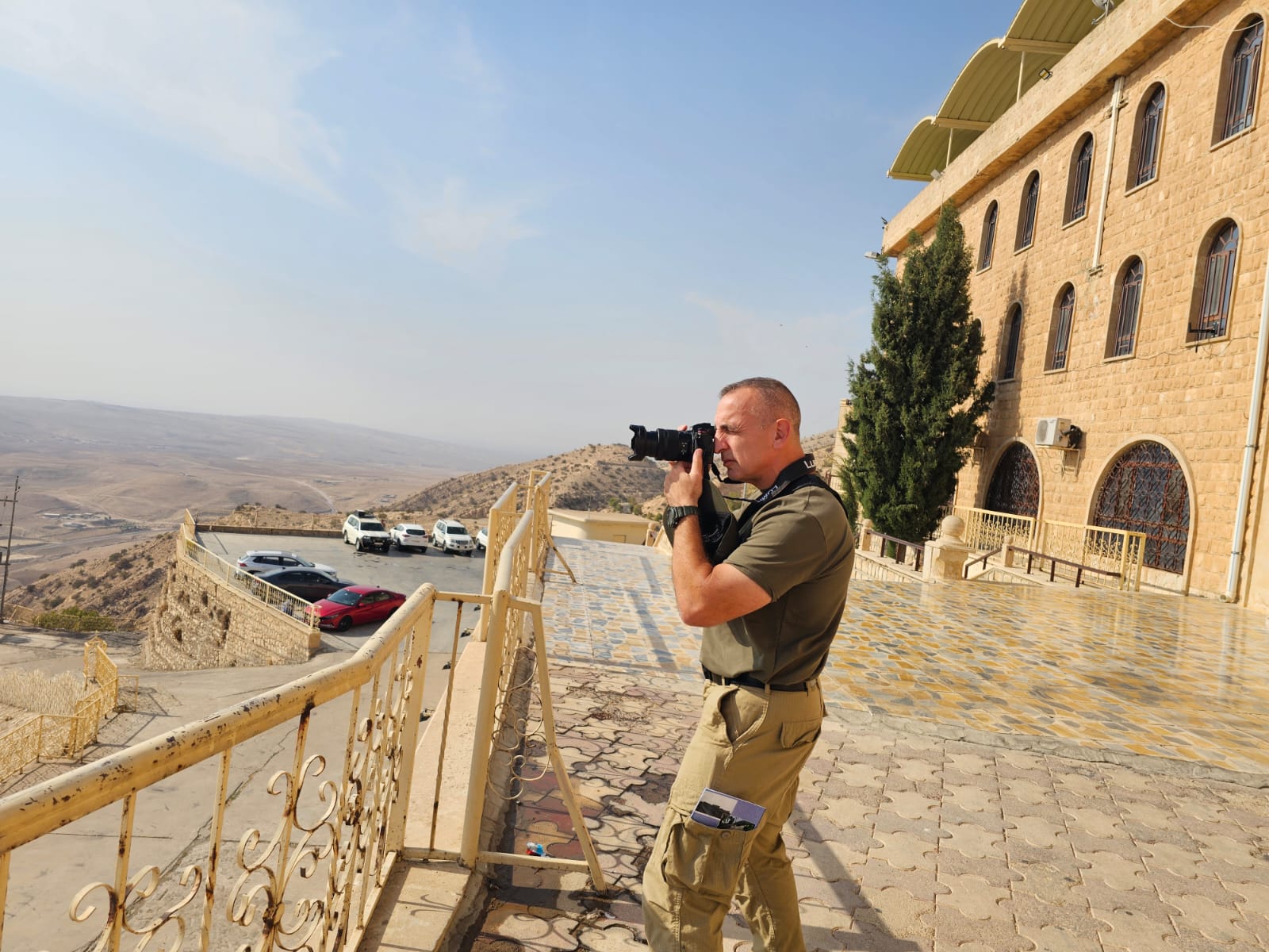 A photographer captures scenic views from a high vantage point near a stone building, surrounded by hills and parked cars.