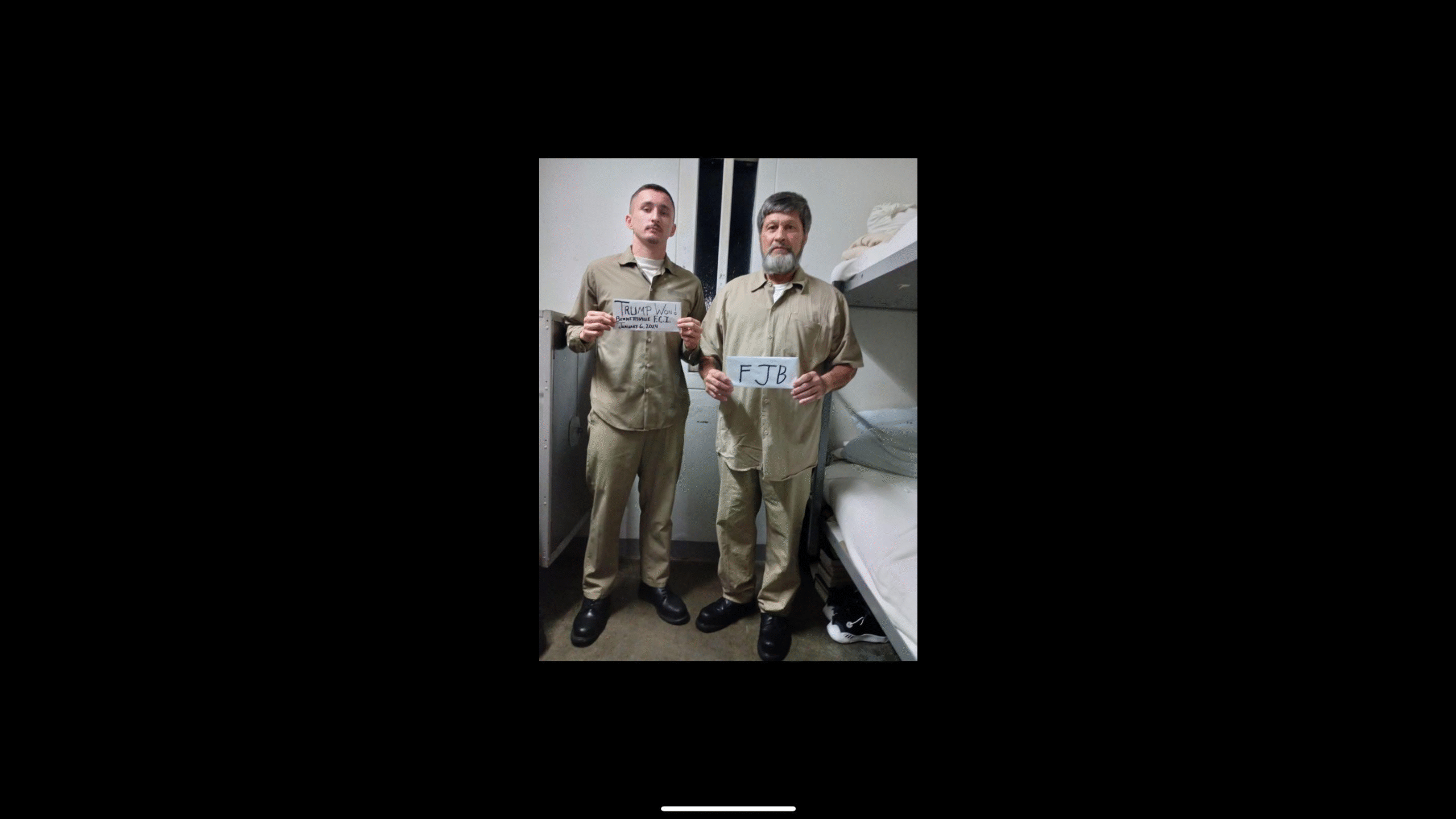 Two men in prison uniforms hold signs with political messages in a jail cell, highlighting a moment of protest or dissent.