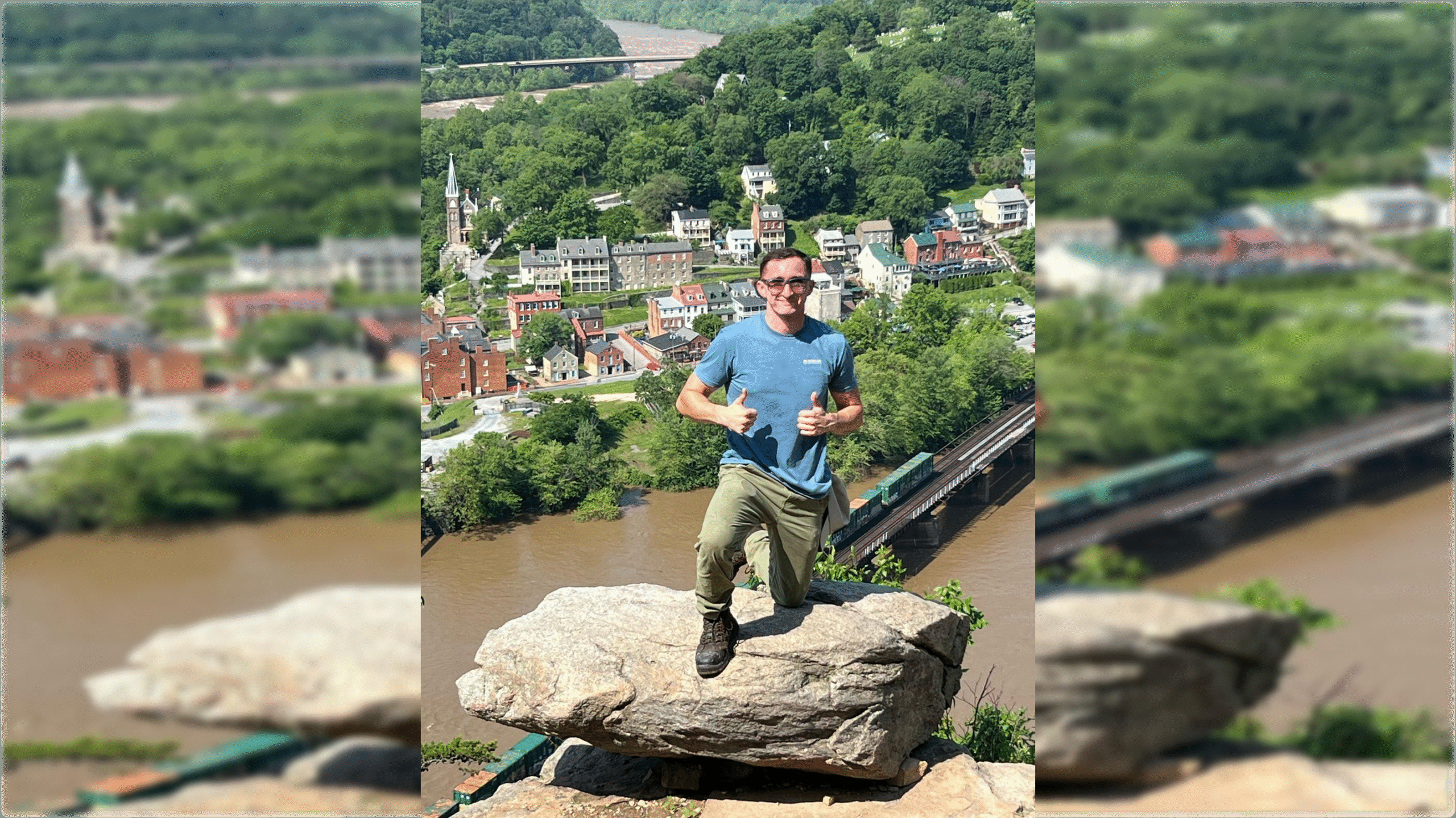 Person kneeling on a large rock with thumbs up, overlooking a scenic view of a town and river surrounded by lush greenery.