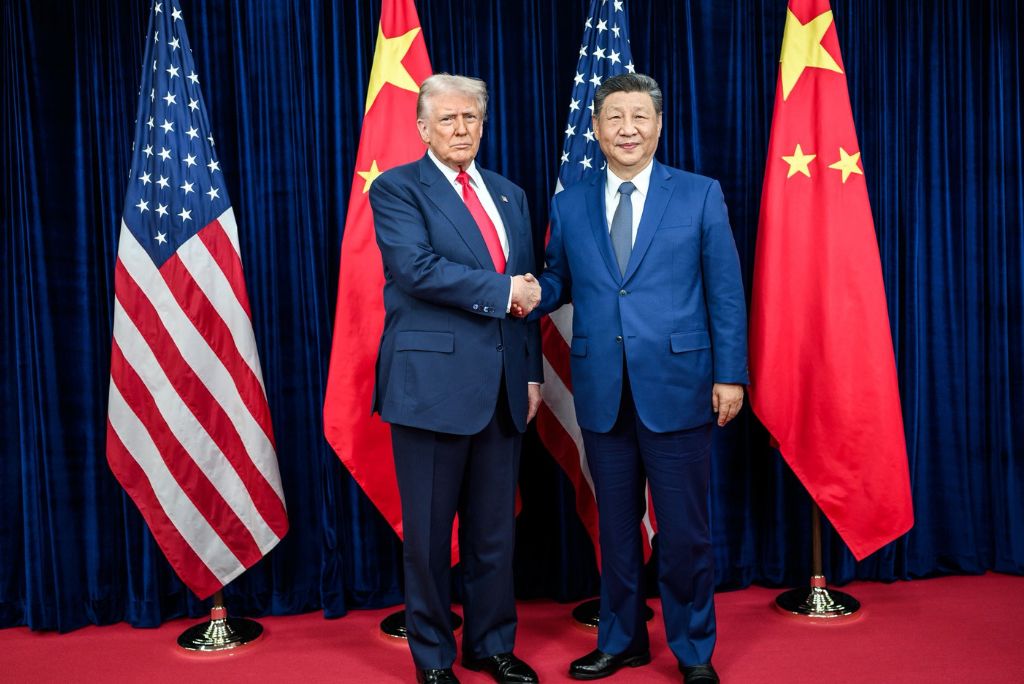 Donald Trump and Xi Jinping shake hands during a diplomatic meeting, flanked by the flags of the United States and China.