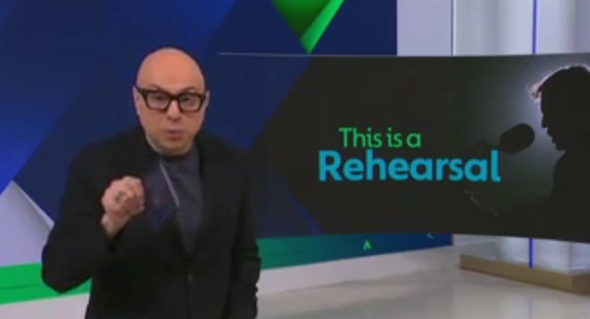 Presenter discussing rehearsal techniques with a graphic displaying the words "This is a Rehearsal" in a studio setting.