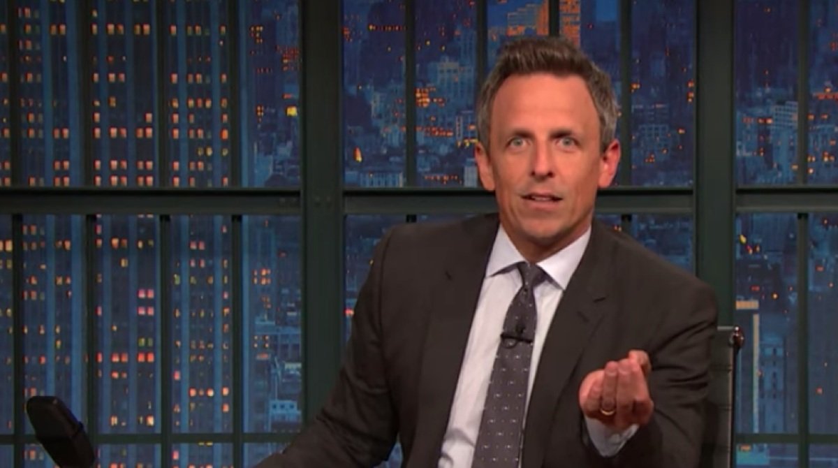 Seth Meyers gesturing while hosting a late-night talk show, with a city skyline illuminated in the background.