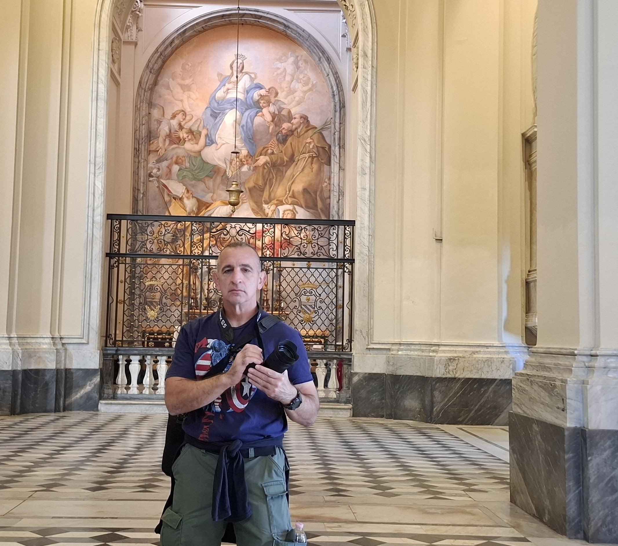 Man holding a camera in a historic building with a large mural and ornate architecture in the background.
