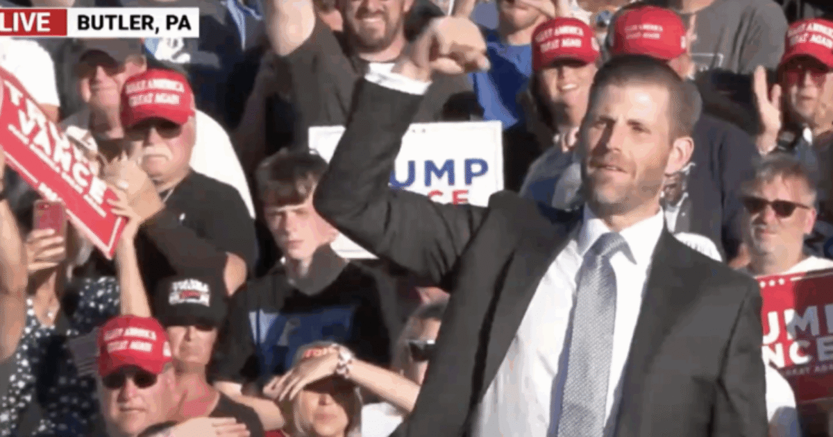 Eric Trump gestures enthusiastically at a rally in Butler, PA, surrounded by supporters wearing red hats and holding campaign signs.