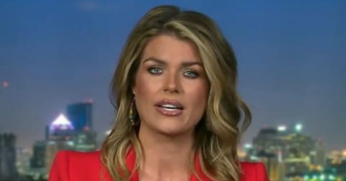 News anchor with long blonde hair wearing a red blazer, presenting against a city skyline backdrop at dusk.