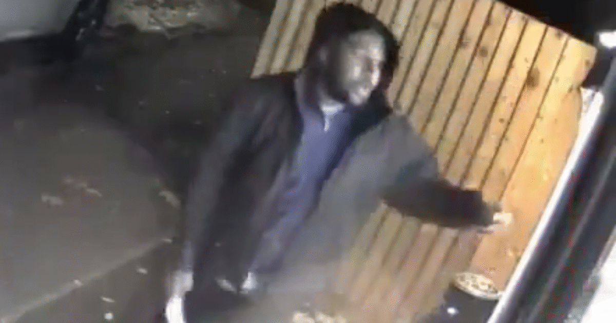 Surveillance image of a man in a black jacket standing near a door, possibly engaging with someone off-camera in an outdoor setting.