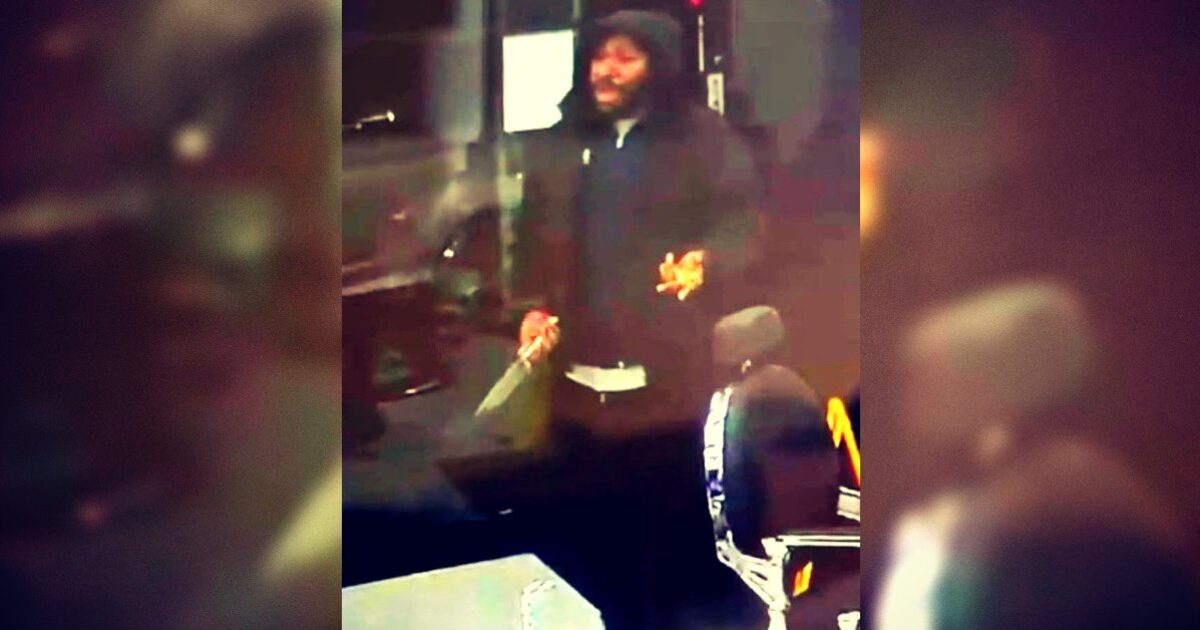 Man holding a knife inside a bus, appearing agitated, with a passenger seated nearby, captured in low-light conditions.