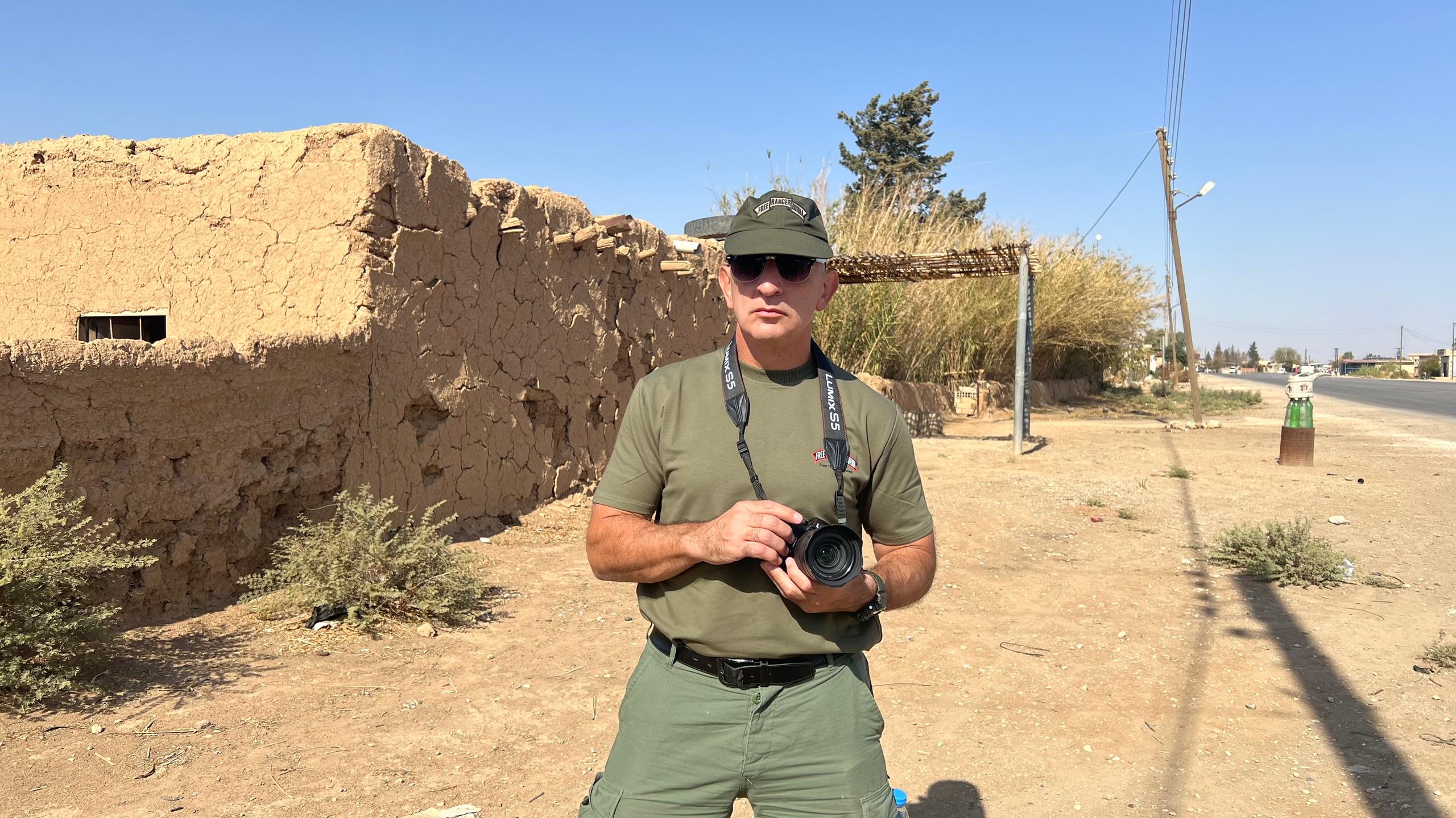 A man in military attire holds a camera in front of a rustic earthen structure under a clear blue sky.
