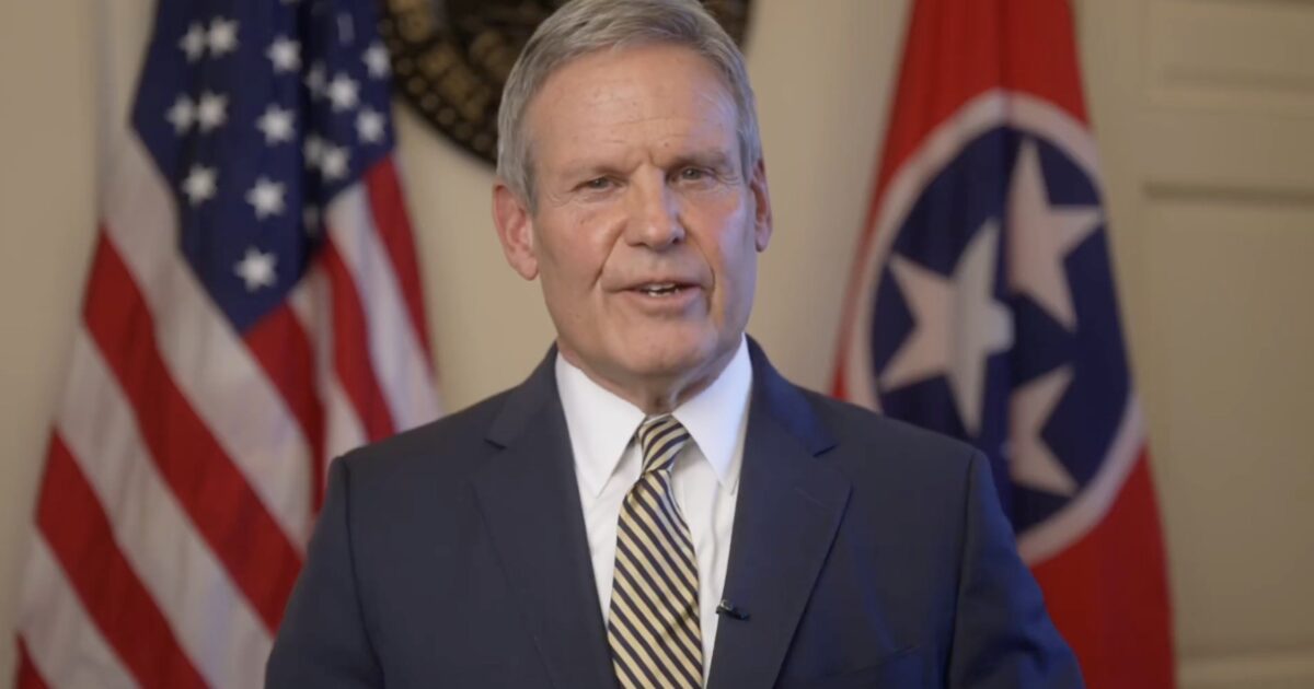 Tennessee governor delivering a message with the state flag and American flags in the background, dressed in a suit and tie.