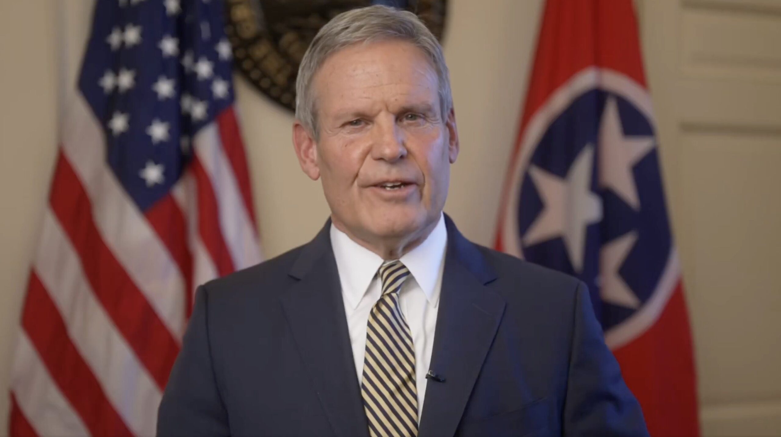 Tennessee governor delivering a message with the state flag and American flags in the background, dressed in a suit and tie.