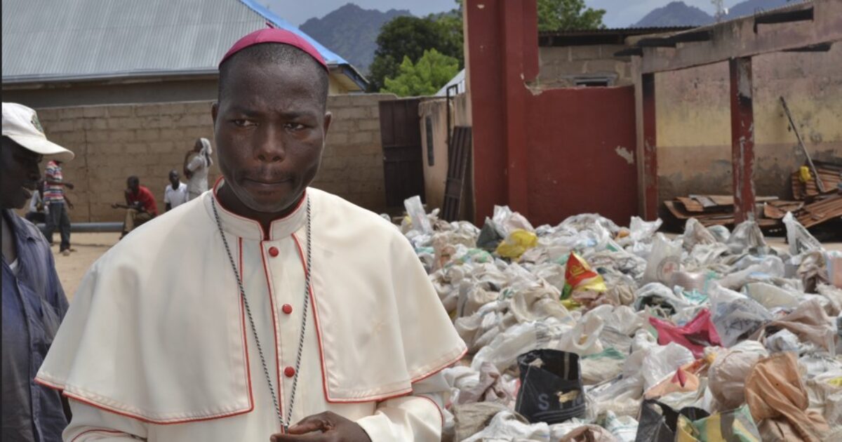 A bishop stands in front of a large pile of discarded materials in a rural community, highlighting environmental issues and waste management challenges.