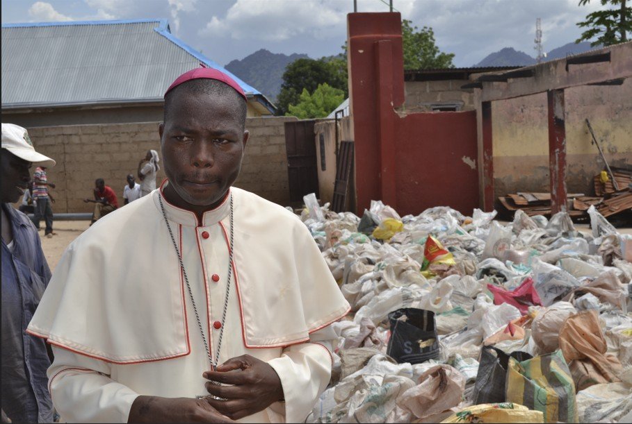 A bishop stands in front of a large pile of discarded materials in a rural community, highlighting environmental issues and waste management challenges.
