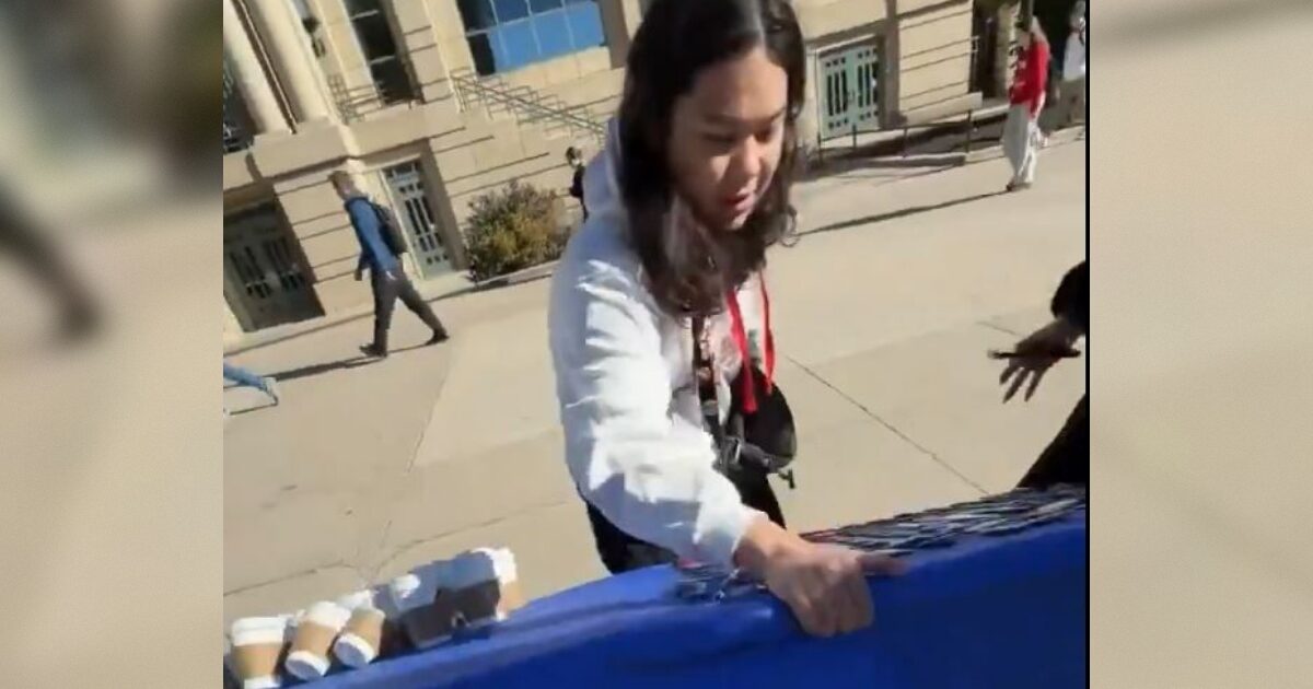 Person setting up a table with items on a sunny day outside a building, with pedestrians walking by in the background.
