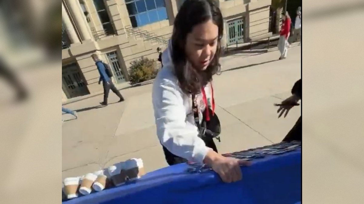 Person setting up a table with items on a sunny day outside a building, with pedestrians walking by in the background.