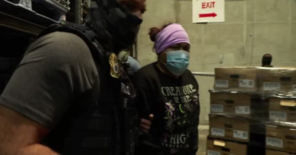 Law enforcement officers escorting a woman wearing a mask and purple headband in a warehouse setting with boxes in the background.