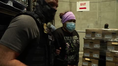 Law enforcement officers escorting a woman wearing a mask and purple headband in a warehouse setting with boxes in the background.