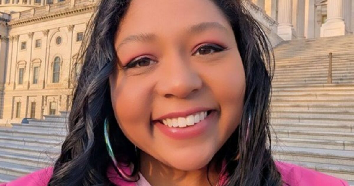 Smiling woman with long dark hair, wearing a pink jacket, stands on steps of a historic building, showcasing a vibrant and cheerful demeanor.