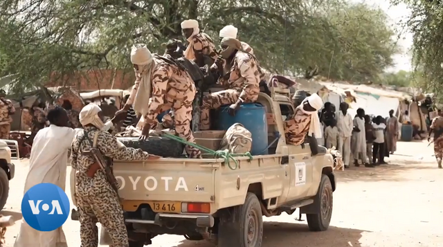 Soldiers in camouflage uniforms load supplies onto a Toyota pickup truck in a desert setting, with people and tents visible in the background.